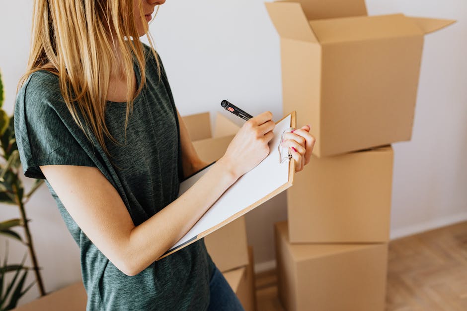 A young woman with long blonde hair, wearing a short-sleeved dark green top, is standing indoors near a wooden floor. She is holding a clipboard with a white sheet of paper and writing with a black marker. Behind her, there are three large, open cardboard boxes stacked on top of each other, placed on the floor. The boxes appear to be empty or partially filled with packaging materials, and their light brown surface has visible corrugated textures. To her left, a potted plant with long, slender green leaves is partially visible. The lighting in the room is soft and natural, suggesting daylight. The scene reflects an environment associated with packing, moving, or preparing for rubbish or waste clearance, aligning with services of independent or private waste handling providers such as Rubbish Removal Richmond, supporting house clearance and related rubbish removal tasks.