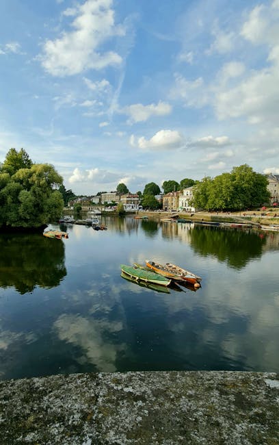 A tranquil river scene with a calm, reflective water surface showing clouds and a blue sky overhead. Several small boats, including a green one and others with brown and wooden finishes, are anchored near the shoreline in the foreground. On the far bank, there are lush green trees and a variety of residential buildings with light-colored facades and dark roofs, set back from the water. The environment appears peaceful and maintained, with no visible rubbish or debris, but the image relates to rubbish removal services through the natural context of waste clearance and environmental management. The scene is well-lit with natural daylight, highlighting the textures of the boats, foliage, and the building facades. The overall composition emphasizes a tidy riverside area, which may be associated with private waste handling and on-site clearance services, as provided by Rubbish Removal Richmond.