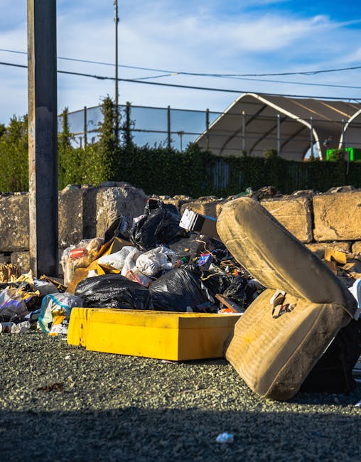 A pile of discarded waste lies on a gravel surface beside a weathered stone wall, with several black and clear plastic rubbish bags mixed with loose debris, including a yellow plastic bin or container. A worn, beige car tyre leans against the pile, partially covering some of the rubbish. In the background, there is a metal pole and overhead power lines, with a row of green bushes and a large, curved, semi-transparent structure—possibly a storage shelter or sports facility—visible against a partly cloudy sky. The scene suggests a waste collection or disposal site, where private rubbish removal services, such as Rubbish Removal Richmond, might handle such bulk waste, highlighting the importance of appropriate on-site clearance and alternative waste handling methods.