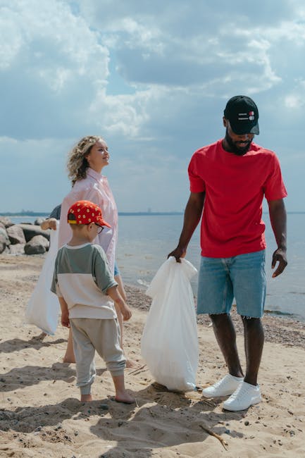 A group of three people, including a man, a woman, and a young boy, stand on a sandy beach near a calm body of water with large rocks along the shoreline. The man, wearing a black cap, red t-shirt, and light blue denim shorts, is holding a large white plastic bag, likely used for rubbish collection or beach clean-up. The young boy, dressed in light grey sweatpants, a light grey t-shirt with short sleeves, and a red cap decorated with yellow spots, is also holding a similar white bag, suggesting participation in an environmental cleanup activity. The woman, with curly blonde hair, is dressed in a white blouse and observing the scene. The sky above features scattered clouds, indicating a partly cloudy day, with natural light illuminating the scene. The background includes the water stretching to the horizon with visible shoreline rocks, creating a setting typical for community efforts in waste disposal and environmental preservation. This image could relate to independent rubbish collection or environmental cleanup initiatives supported by waste management services like Rubbish Removal Richmond.