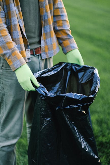 A pile of discarded waste lies on a gravel surface beside a weathered stone wall, with several black and clear plastic rubbish bags mixed with loose debris, including a yellow plastic bin or container. A worn, beige car tyre leans against the pile, partially covering some of the rubbish. In the background, there is a metal pole and overhead power lines, with a row of green bushes and a large, curved, semi-transparent structure—possibly a storage shelter or sports facility—visible against a partly cloudy sky. The scene suggests a waste collection or disposal site, where private rubbish removal services, such as Rubbish Removal Richmond, might handle such bulk waste, highlighting the importance of appropriate on-site clearance and alternative waste handling methods.
